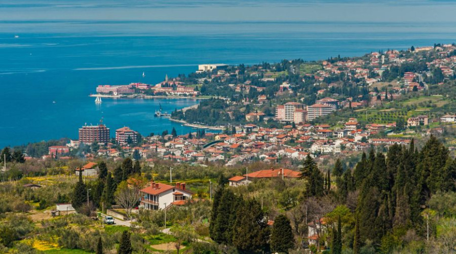 Panoramski pogled na obalno mesto Portorož z zgradbami in zelenjem, ki se raztezajo nad turkiznim morjem.