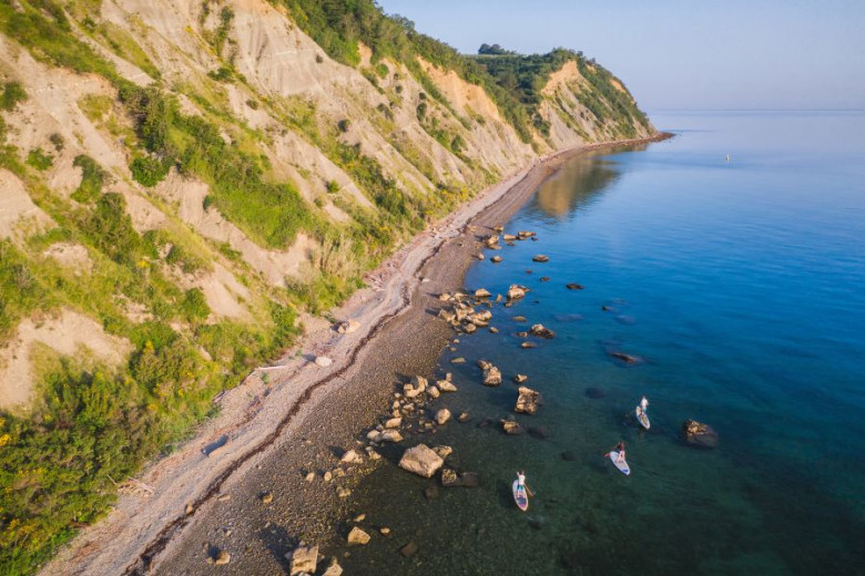 Pogled iz zraka na štiri ljudi na supih v čistem morju blizu skalnate plaže ob vznožju strmega, z vegetacijo poraslega klifa.