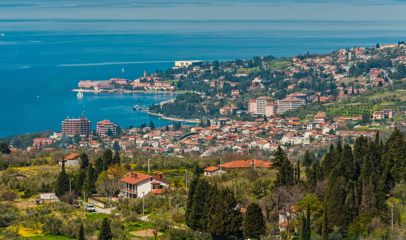 Panoramic view of the coastal town of Portorož, with buildings and greenery stretching above the turquoise sea.