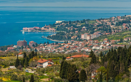 Panoramski pogled na obalno mesto Portorož z zgradbami in zelenjem, ki se raztezajo nad turkiznim morjem.