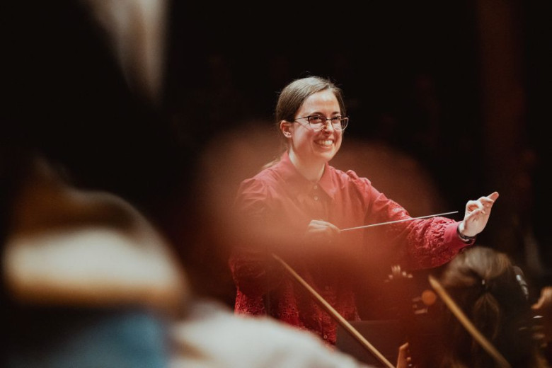Smiling Mojca Lavrenčič leads an orchestra, she is holding a baton in her hand.
