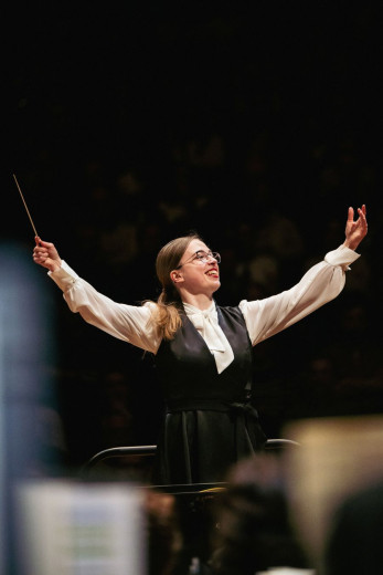 Conductor Mojca Lavrenčič stands on stage with her arms outstretched, holding a baton in one hand.