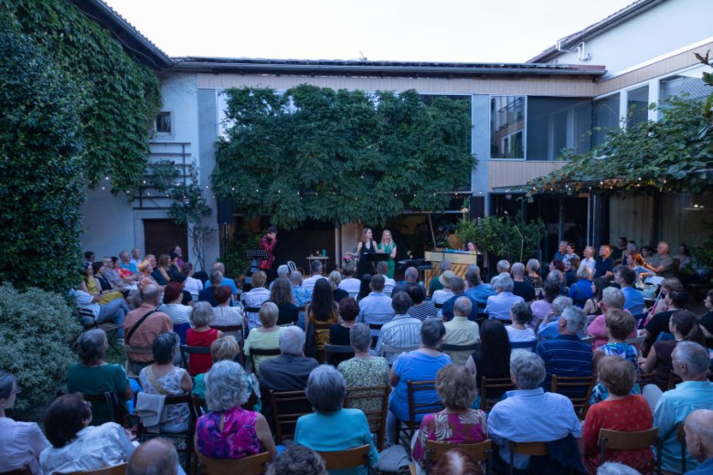 Outdoor event with a large audience seated in a lush, green courtyard. Two people perform on a stage.