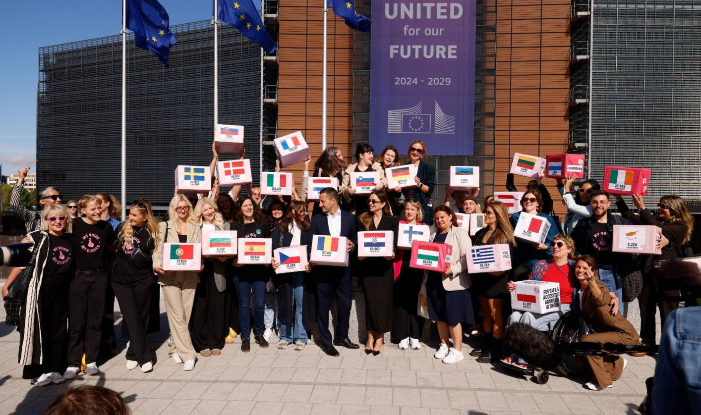 People are standing outside of a building and taking a group photo. Each is holding a cube with a different European flag on it. Three EU flags are waving on poles in the background.