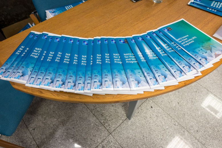 Several copies of a blue brochure titled "NATO Science for Peace and Security (SPS) Programme" arranged on a circular wooden table, adjacent to a blue chair.