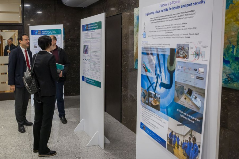 Three individuals are viewing research posters displayed on white stands. The posters contain text, graphs, and scientific illustrations.