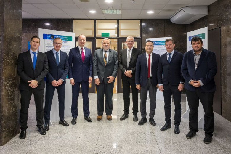 Nine men in business attire standing shoulder to shoulder and posing in front of two informational posters. They are smiling directly at the camera.
