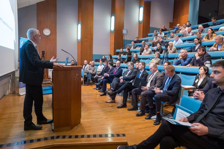 Auditorium view of a speaker at a podium addressing an audience, who are seated in tiered rows.