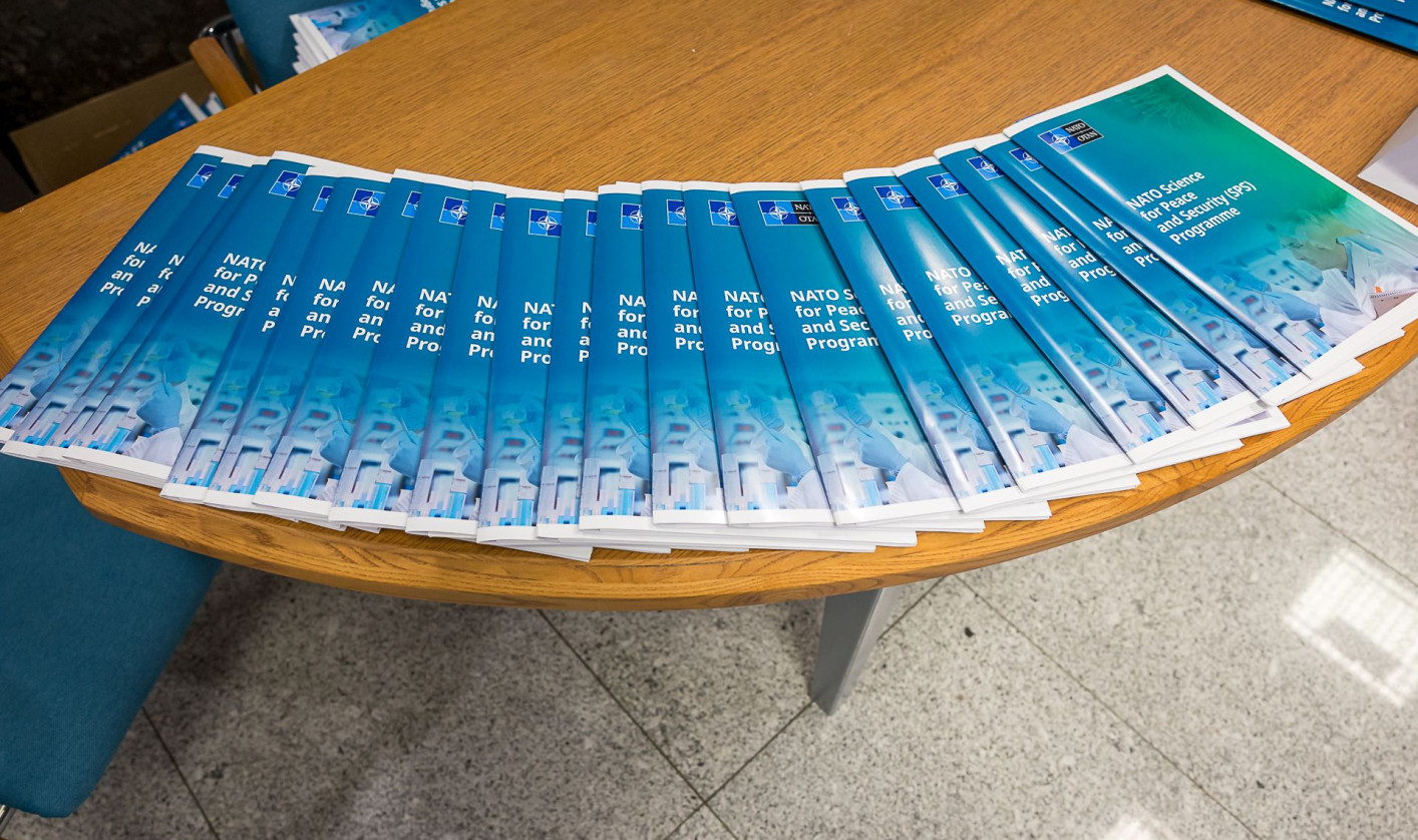Several copies of a blue brochure titled "NATO Science for Peace and Security (SPS) Programme" arranged on a circular wooden table, adjacent to a blue chair.