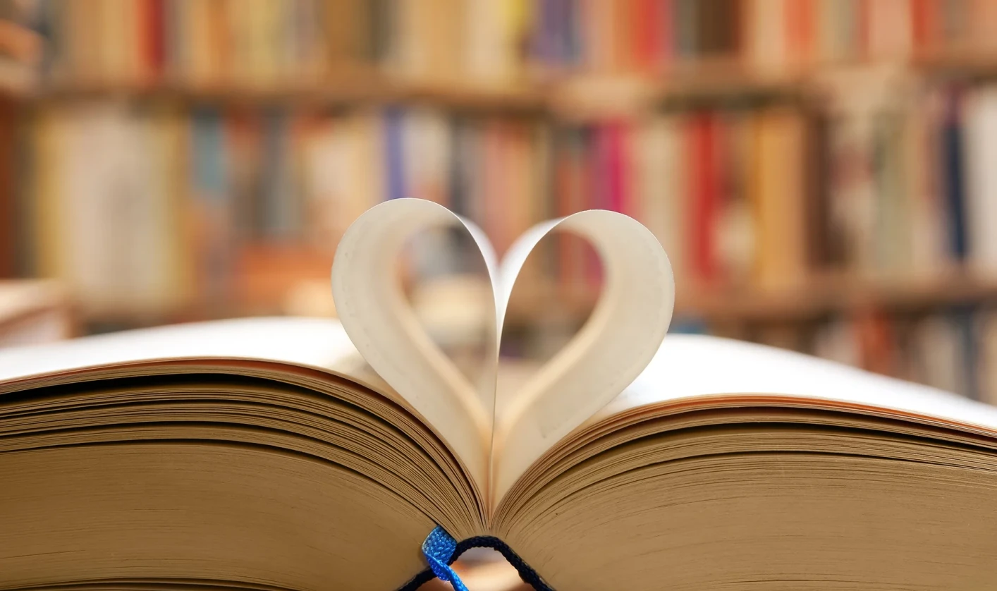Open book with pages shaped into a heart in front of a bookshelf in the background.