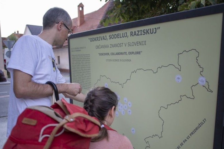 The image shows a man and a girl looking at an information board titled ‘Discover, Collaborate, Explore – Citizen Science in Slovenia.’ The board features a map of Slovenia with several marked locations. The man, wearing glasses and a white T-shirt, point