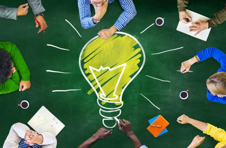 A group of people sitting around a green table, viewed from above. In the center of the table, there is a large drawing of a yellow light bulb with white rays, symbolizing creativity and ideas. 
