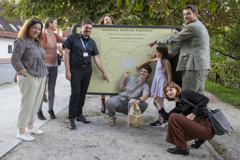 A group of nine people standing and posing next to a large information board titled ‘Discover, Collaborate, Explore – Citizen Science in Slovenia.’ The board displays a map of Slovenia with several marked locations. 