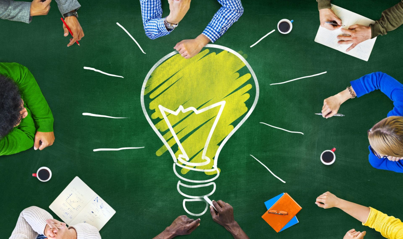 A group of people sitting around a green table, viewed from above. In the center of the table, there is a large drawing of a yellow light bulb with white rays, symbolizing creativity and ideas. 
