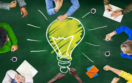 A group of people sitting around a green table, viewed from above. In the center of the table, there is a large drawing of a yellow light bulb with white rays, symbolizing creativity and ideas. 