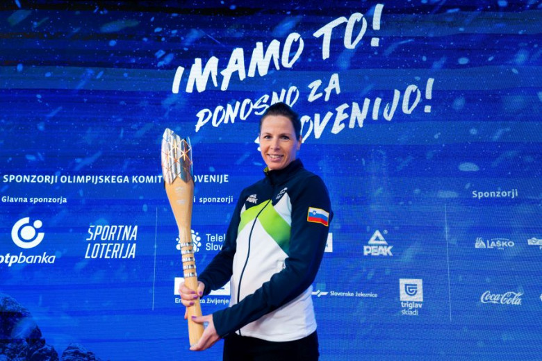 Ženska v športni jakni drži zlato olimpijsko baklo in se smehlja pred modrim ozadjem z logotipi sponzorjev.