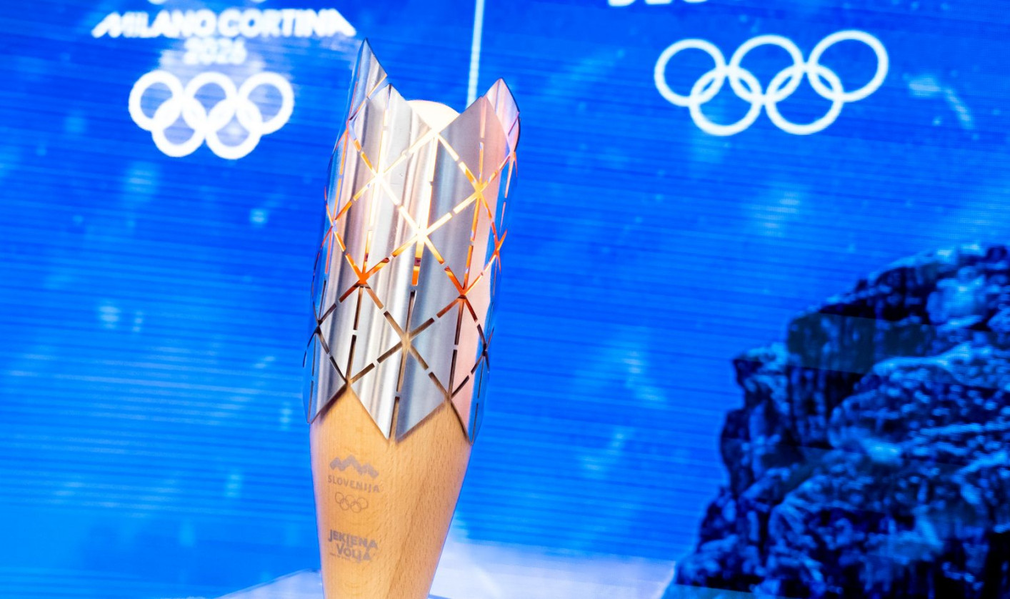 A golden Olympic torch stands upright, its patterned metal top glowing against a blue background with Slovenia and Olympic logos.