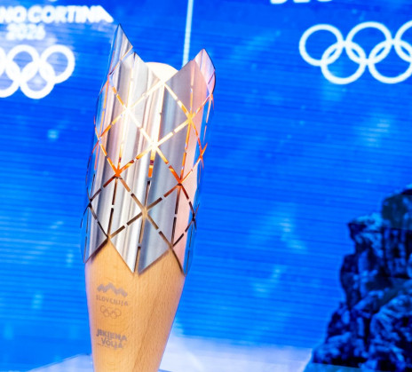 A golden Olympic torch stands upright, its patterned metal top glowing against a blue background with Slovenia and Olympic logos.