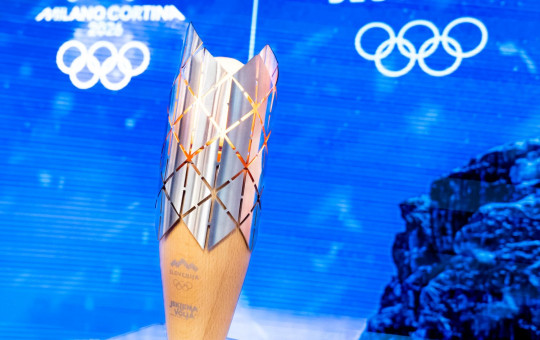 Zlata olimpijska bakla stoji pokončno, njen kovinski vrh z vzorcem izstopa pred modrim ozadjem z olimpijskimi simboli.