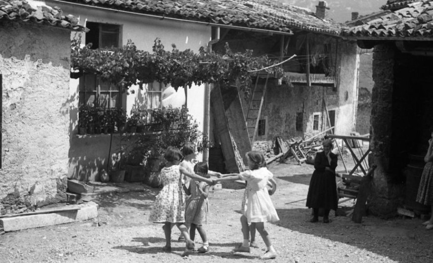 Children dance in a circle in an old village courtyard as an older woman watches from a distance.
