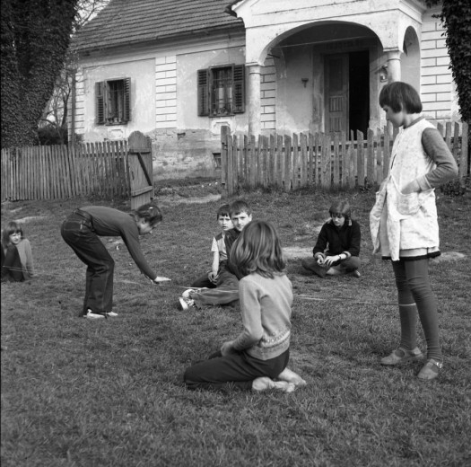 Children play a rubber-band jumping game in a grassy yard, while others watch nearby.