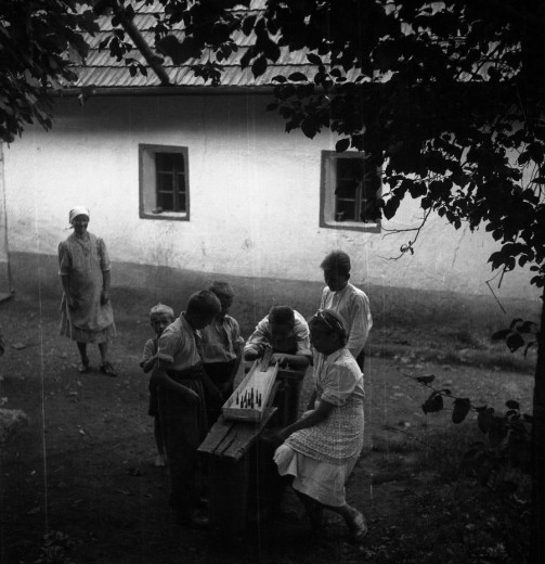 Children and women gather by a bench in front of a house as the kids play a board game with wooden pieces.
