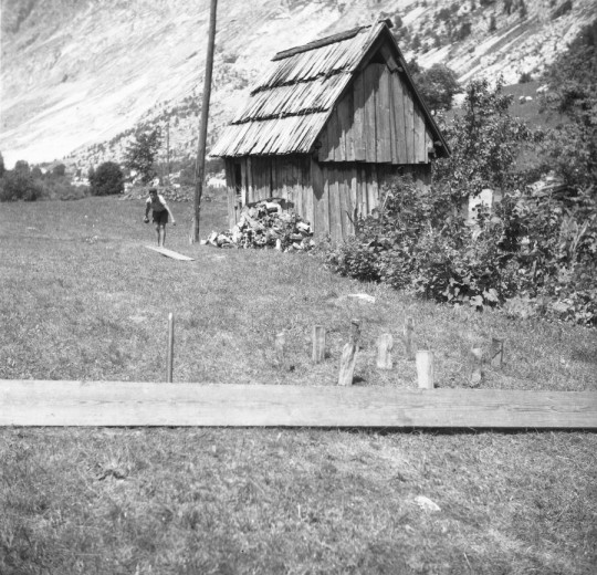 A simple skittle game stands on the meadow before an old wooden hut, with a child walking in the background.
