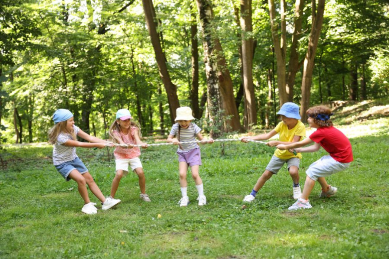 Children play a teamwork tug-of-war game on the grass in a forest clearing.