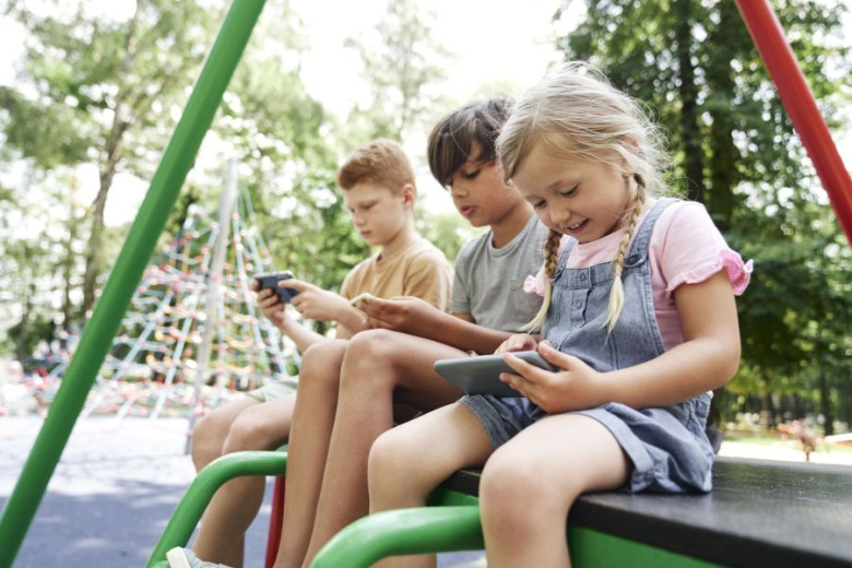 Children on a playground sit together while looking at their screens of mobile phones.