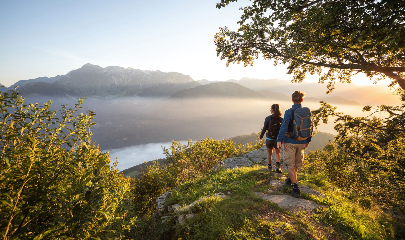 Two people with backpacks stand on a grassy viewpoint overlooking misty mountains and a valley at sunrise.