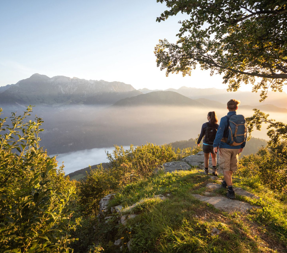 Dva človeka z nahrbtniki stojita na travnati razgledni točki s pogledom na megleno hribovje in dolino ob sončnem vzhodu.