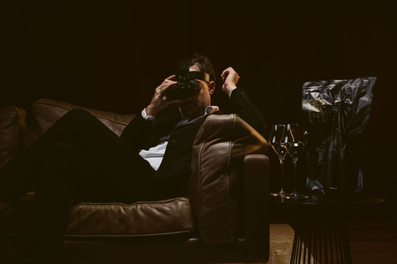Moški v črnem sedi v temi na rjavem kavču in gleda skozi daljnogled v popolni temi