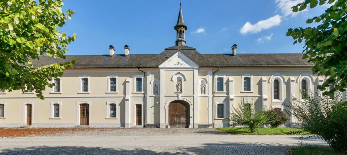 Zgodovinska samostanska fasada z bež stenami in obokanimi lesenimi vrati pod jasnim modrim nebom. Listnata drevesa uokvirjajo miren prizor na obeh straneh.