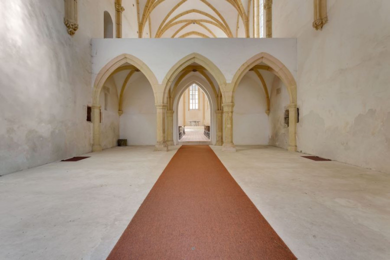 Zgodovinska notranjost cerkve z visokim obokanim stropom in kremnimi stenami. Rdeča preproga poteka po sredini proti svetlemu oltarju in izraža spokojnost.