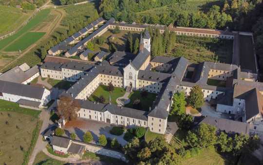 Aerial view of a large, historic monastery complex surrounded by lush greenery and fields, with a central courtyard and a serene, peaceful ambiance.