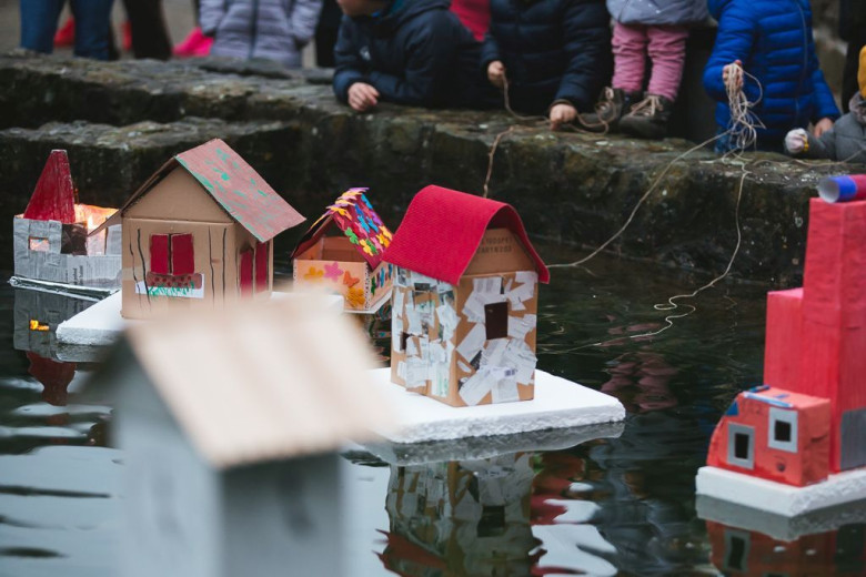 Otroci so zbrani ob ribniku in opazujejo barvite papirnate modele hiš, ki plavajo na stiroporju. Prizor je igriv in ustvarjalen.