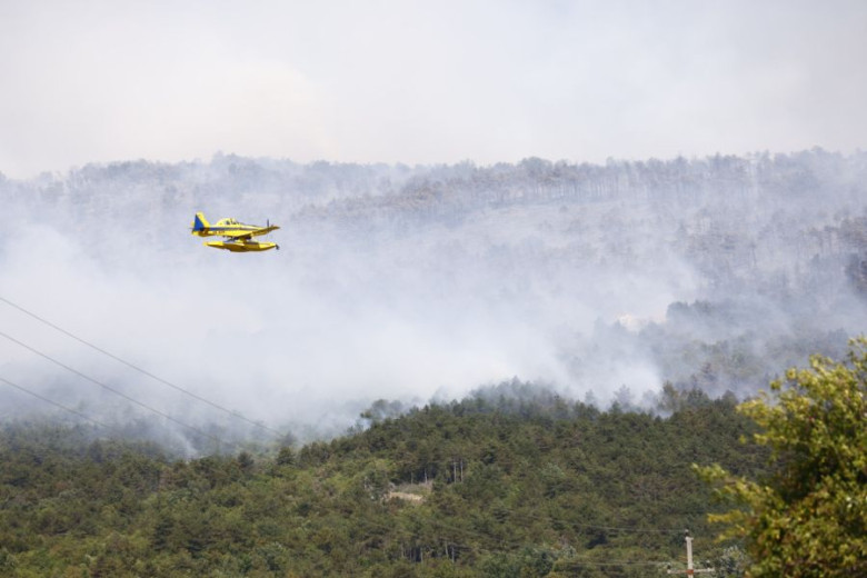 A firefighting plane is flying over the hill engulfed in flames.