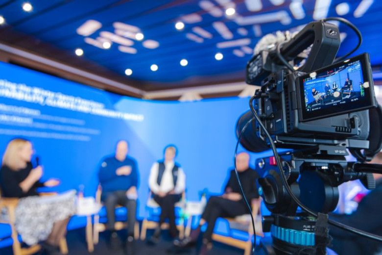 Panelists sitting on stage being filmed on camera.