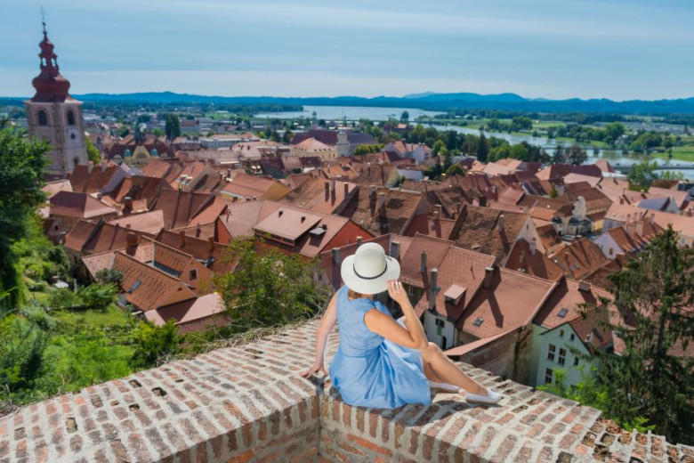 Woman sitting on a viewpoint, looking over a historic town with towers and distant hills.