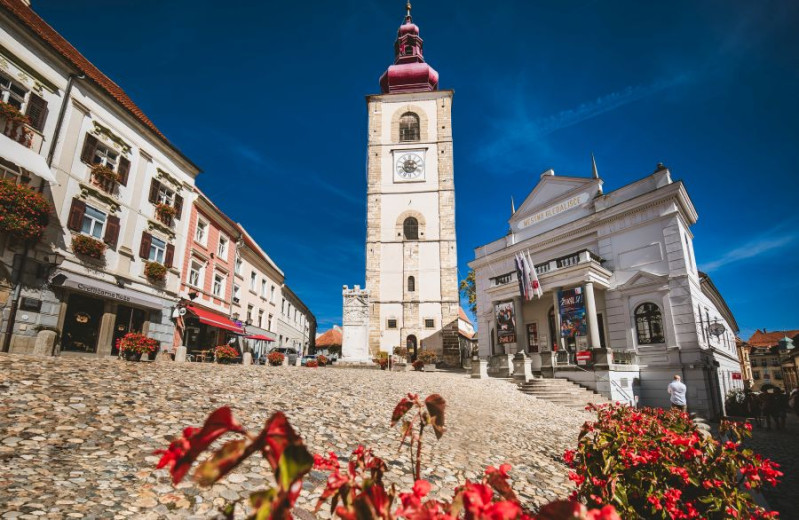 Sunlit historic square with clock tower, theatre, and colorful flowers.