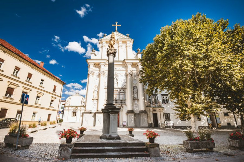 A square with a tall column, a historic church, surrounding buildings, and shaded seating.