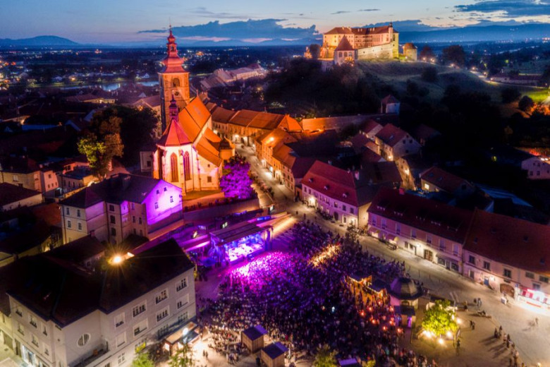 Nighttime view of a lively town festival with lights, music, and crowds beneath the hilltop castle.