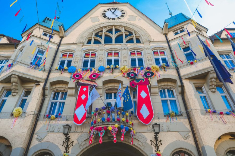 The building is decorated with iconic Kurent costume elements—bright ribboned masks, long red tongues, feathers, and festive ornaments that echo the traditional fur-covered figure.