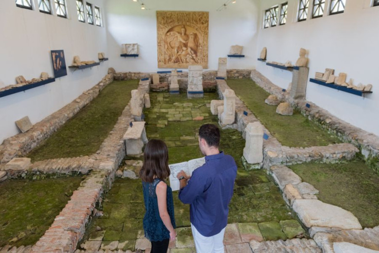 Two visitors exploring an indoor archaeological site with preserved Roman remains and stone artifacts.
