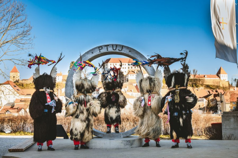 Kurents stand before a large ring-shaped sculpture marked Ptuj, with the town and castle rising in the background as they showcase their traditional costumes.