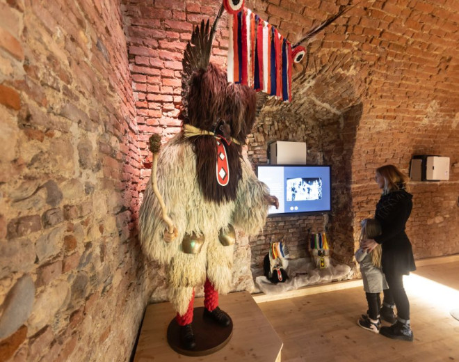 A child and adult view a full Kurent costume on display, with its tall feathers, colorful ribbons, fur suit, and cowbells showcased in a historic brick room.