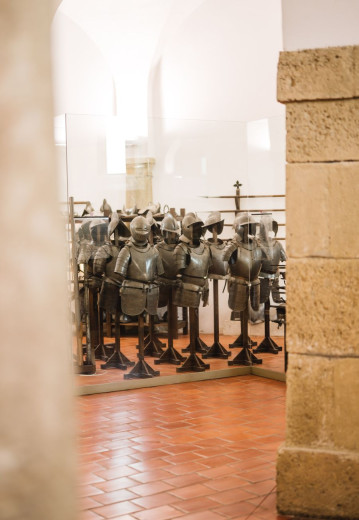 A display of medieval armor suits arranged in rows inside a historic museum room with stone walls and tiled floors.