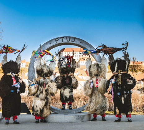 Kurents stand before a large ring-shaped sculpture marked Ptuj, with the town and castle rising in the background as they showcase their traditional costumes.