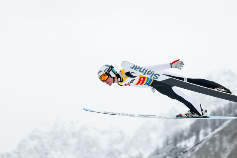 Ski jumper Domen Prevc in mid-air during a jump, wearing a helmet and ski suit, with snowy mountains in the background. 