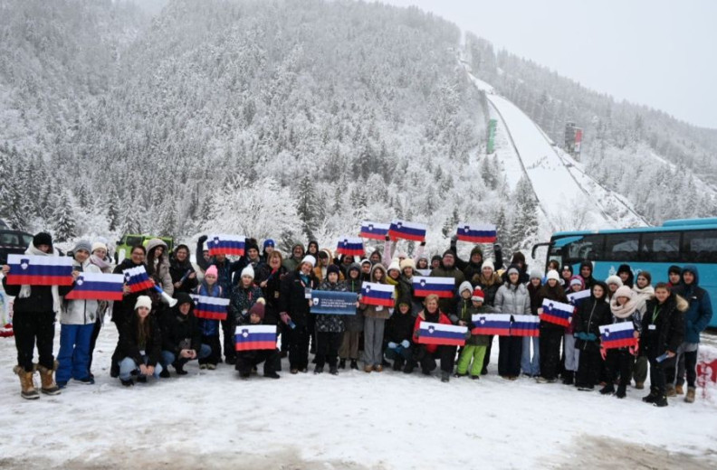 A group of smiling young people holding Slovenian flags pose for a photo in front of the ski jumping hill in Planica.